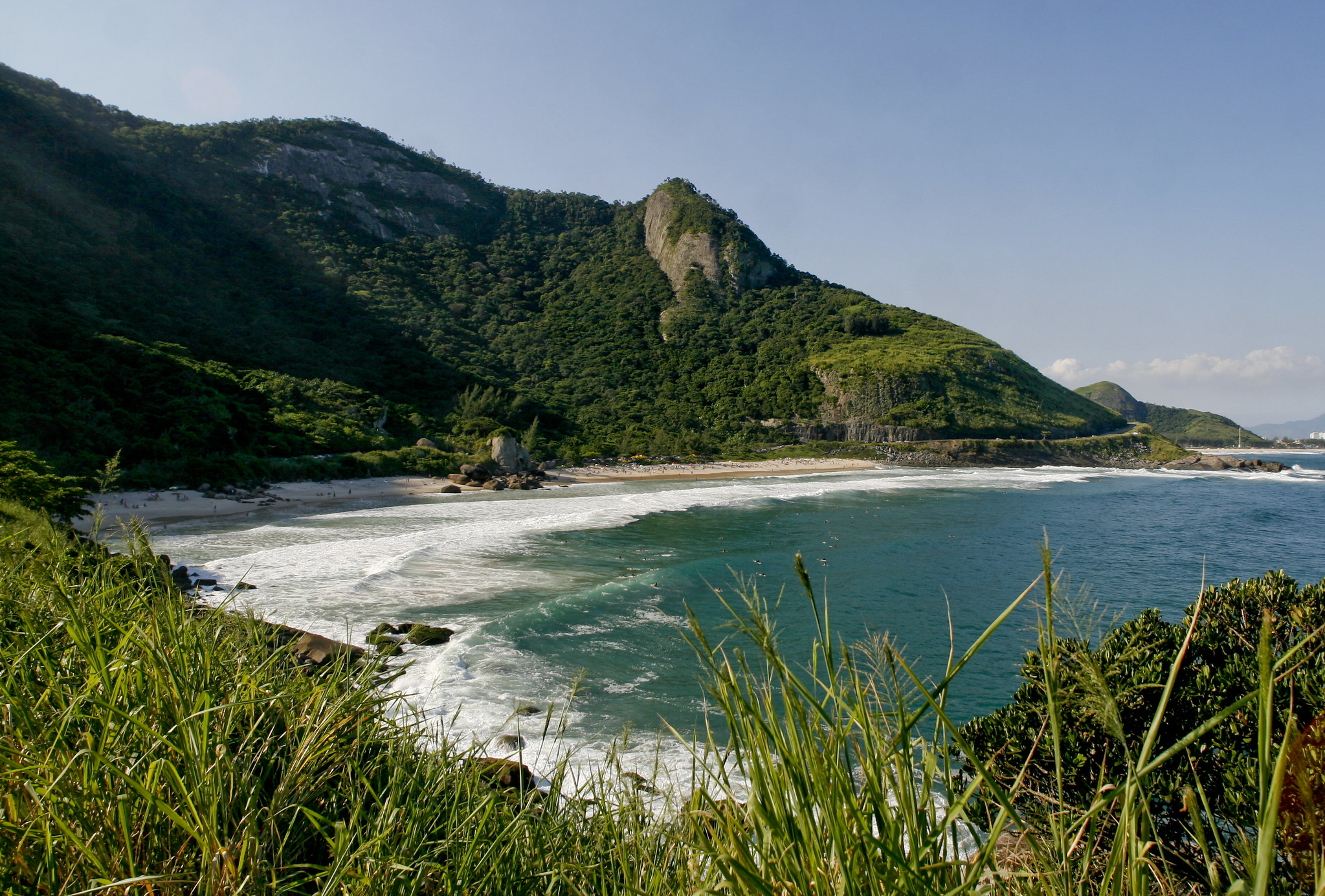 A nearly empty crescent beach wraps around a bay enclosed by forested hills. Foamy waves gently lap the sand. Some people appear to be gathered towards the far end of the beach. Image: Pedro Kirilos/Riotur