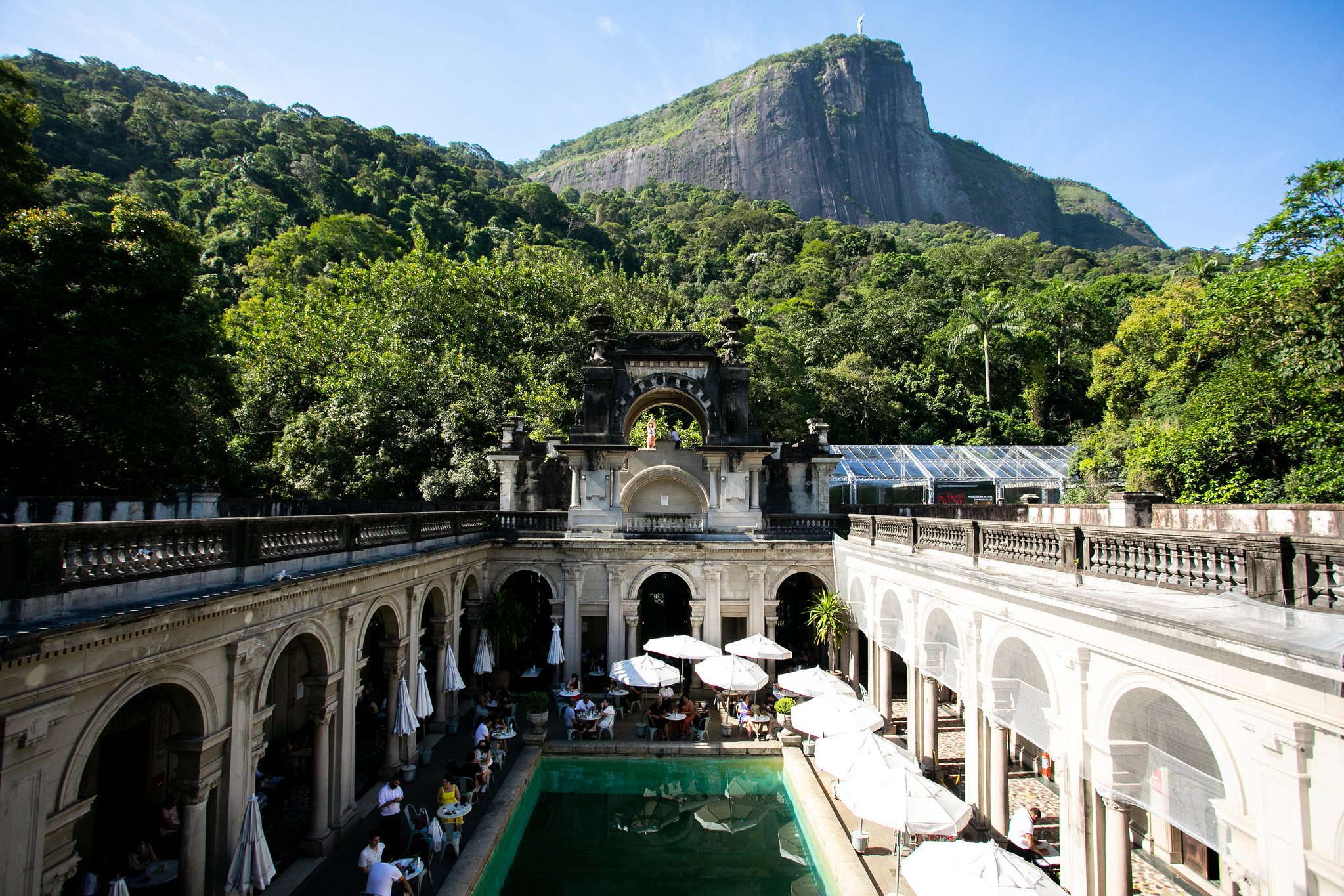 A long, narrow stone quadrangle with a pool at the centre. The pool is surrounded by a low stone wall. Three sides of the quadrangle are visible, each made of a series of arches that open onto a covered, tiled walkway. Between the pool and the walkways, tables are set out. People are seated at these tables and appear to be dining. Leafy hills are visible behind the quadrangle and, in the far distance, Corcovado mountain is visible, with Christ the Redeemer perched atop it.