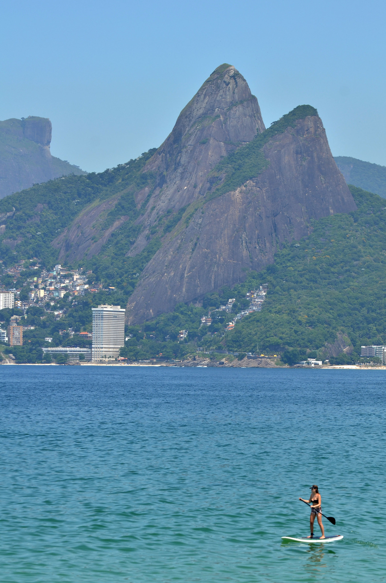 A stretch of mountainous coastline with a cluster of buildings at the point where the mountains meet the sea. A figure paddleboards on the sea in the foreground. They appear tiny compared to the mountains and the sea. Image: Alexandre Macieira/Riotur