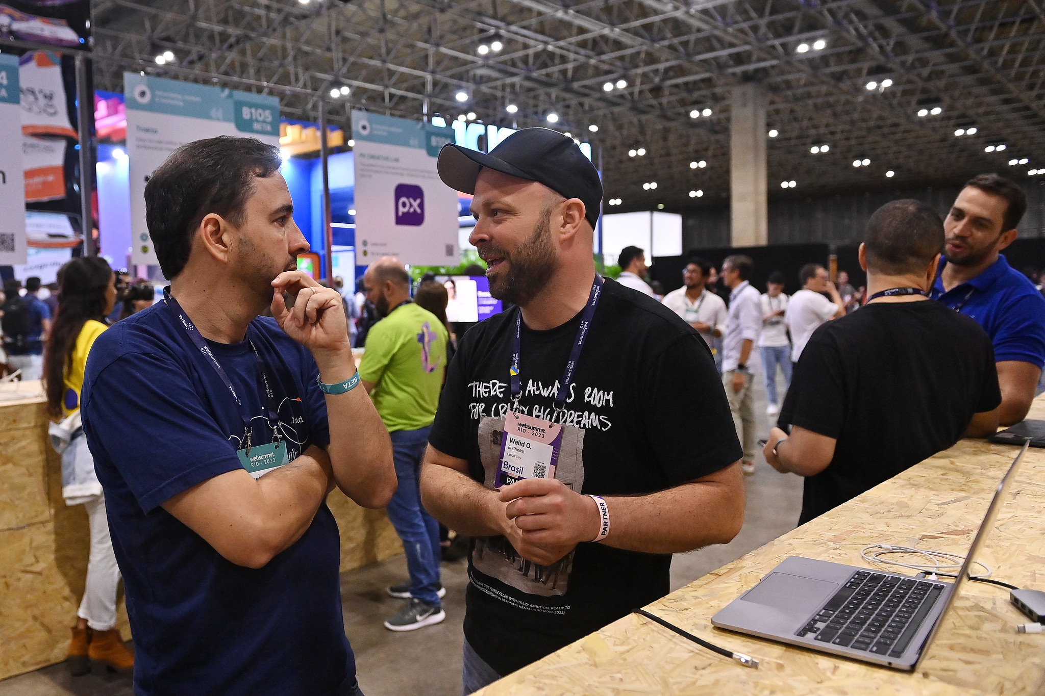 Two people in conversation. The person who appears to be speaking is wearing a baseball cap and a crew-neck tshirt, which reads 'There's always room for crazy big dreams'. This person is gesturing with their left hand, and their right arm is crossed in front of their stomach. The second person, also wearing a crew-neck tshirt, has their arms folded over their chest, with one hand raised towards their face. They appear to be listening attentively.
