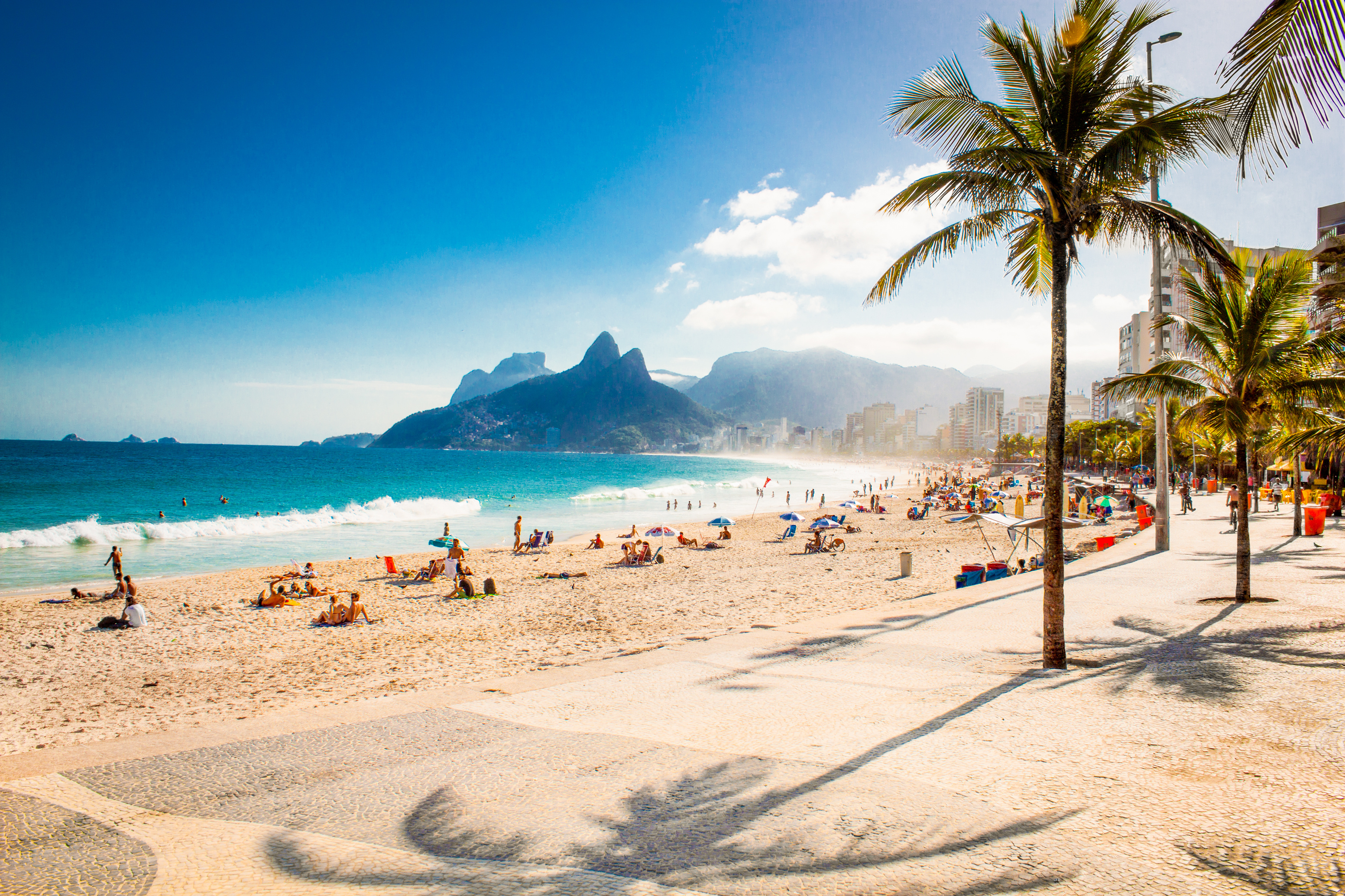 A beach with people lying in small groups on the sand. It's bright, and the palm trees in the foreground are casting shadows. A small wave ripples the sea. There are mountains in the background.