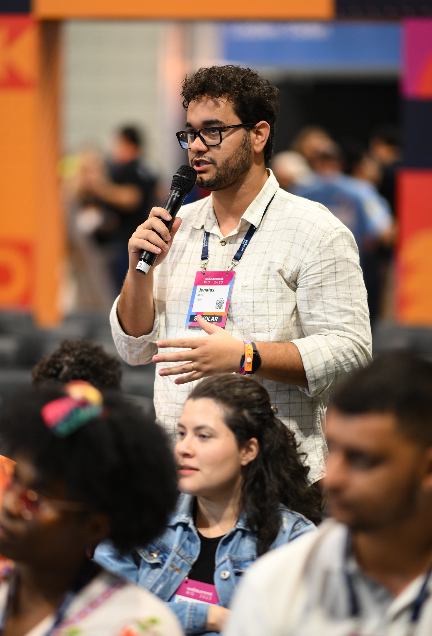 A person stands in the middle of a seated audience. The person is facing the same way as the audience. They are holding a microphone in their right hand and gesturing with their left. They appear to be speaking to someone out of shot.