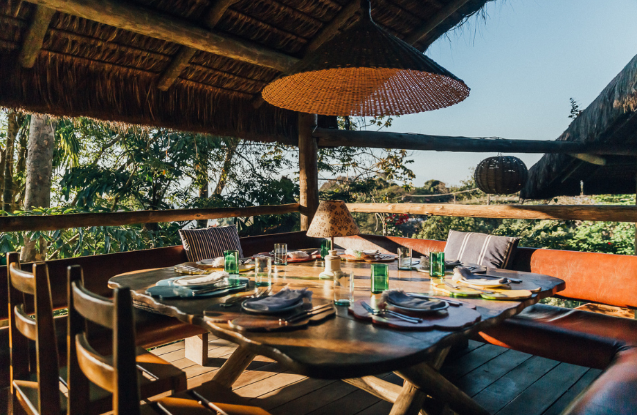 A wooden table set for a meal, with glasses, plates, cutlery and linen. Wooden chairs and benches are pulled up to the table. A wicker lampshade is suspended above the table, and is hanging from a straw roof. The table is surrounded by vegetation.