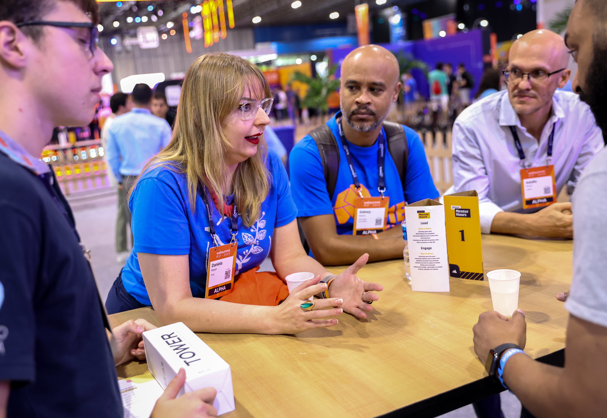 A person leans their elbows on a standing table. They're gesturing with their hands, and appear to be speaking. Two people to their left are looking towards them and appear to be listening. These two people are also leaning on the table.