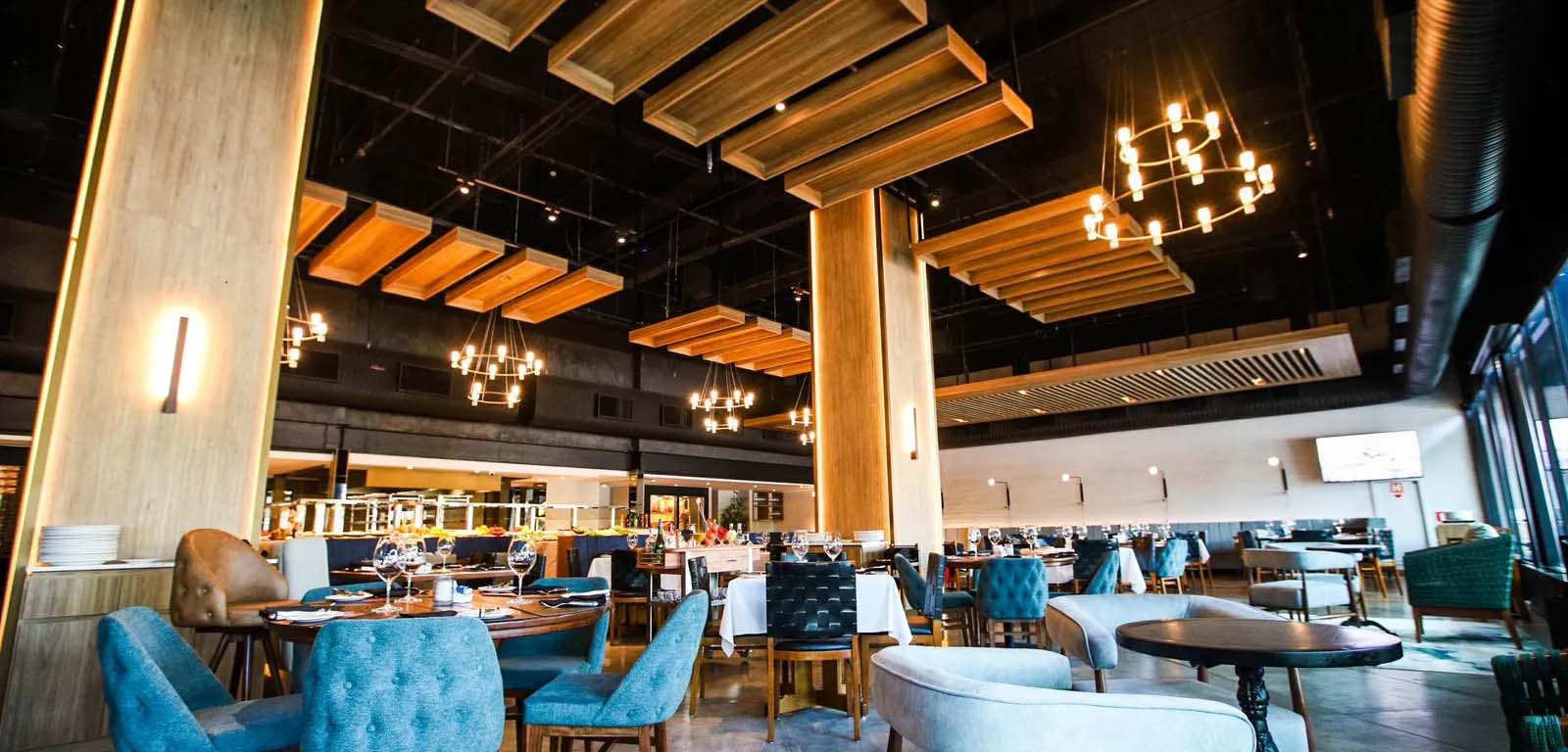 The interior of a restaurant. This is an industrial space, with round tables and what appear to cloth-covered chairs dotted around a cement floor. Two large, square pillars support the ceiling. Lighting and wooden panels are sparsely suspended from the ceiling. The kitchen is open to the restaurant and is visible in the background.