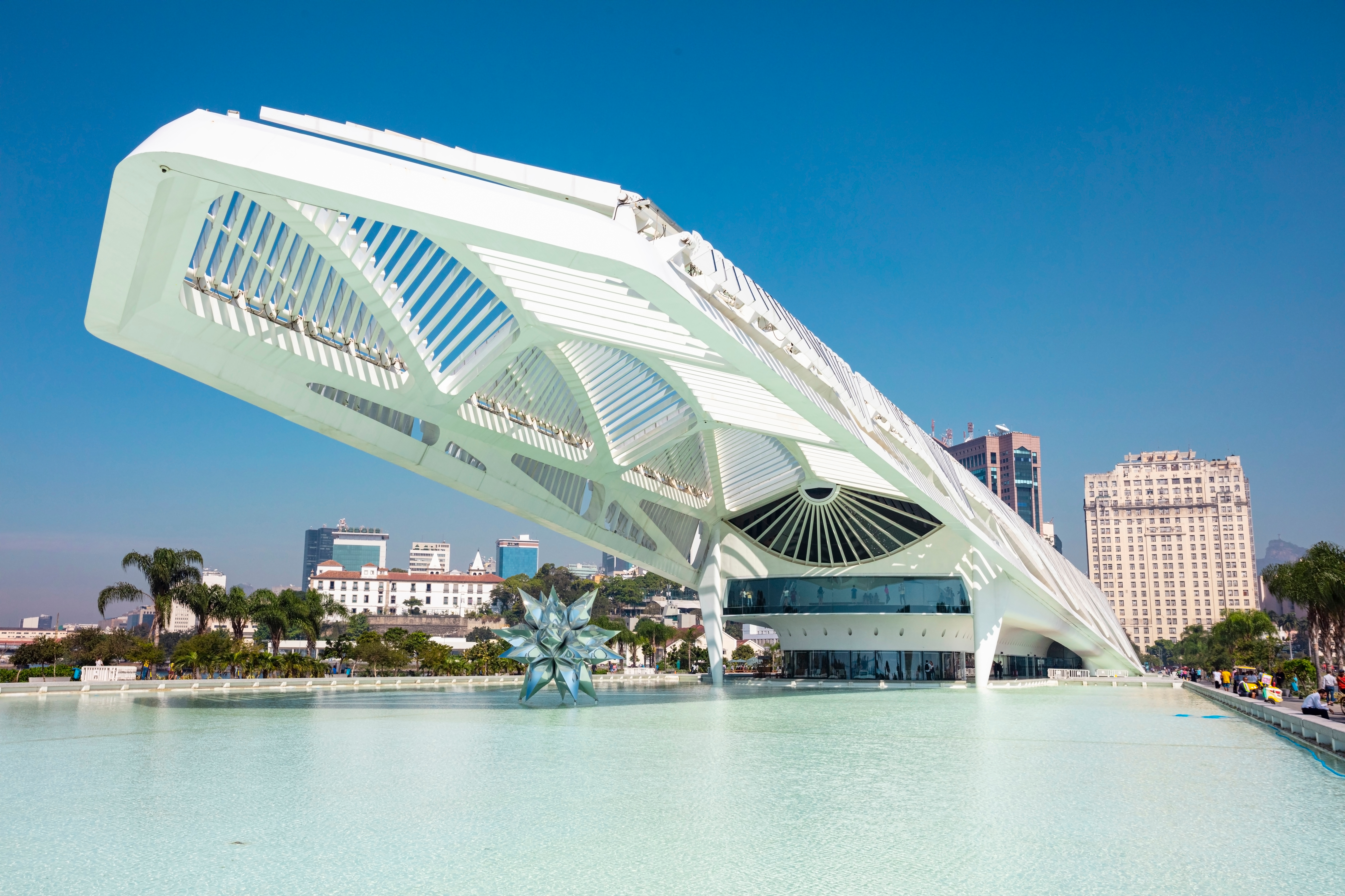 A modern building beside a reflecting pool. The building’s roof extends from the front of the building over the pool. This extended section of roof is made up of dozens of narrow panels that appear to be attached to the structure with hinges. Under the roof, standing in the pool, is a sculpture of a 20-point star. Image: wtondossantos/Shutterstock