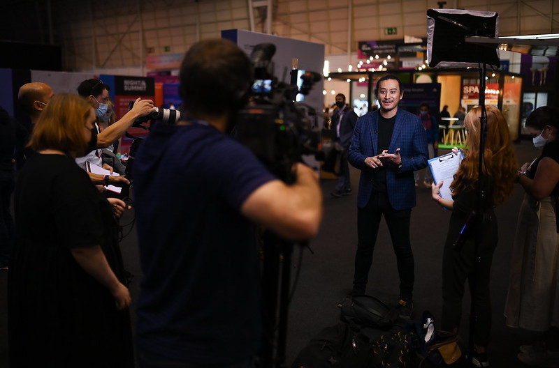 A person (Junta Nakai of Databricks) stands on what appears to be an exhibition floor. Several large stands and lounge areas are visible behind Junta. A group of people stand in front of Junta, facing Junta – some with clipboards and others with cameras. Junta is looking towards a video camera. Junta appears to be taking part in an interview.