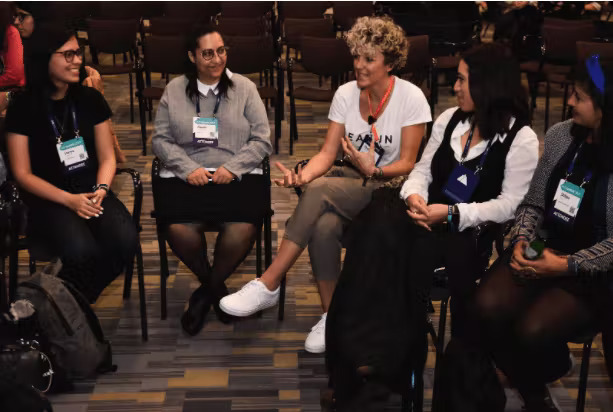 A group of five people sit together. They appear to be a segment of a circle of people. The person in the center is gesturing with their right hand and appears to be speaking, and the other four – who each have their hands clasped in their laps – appear to be listening.