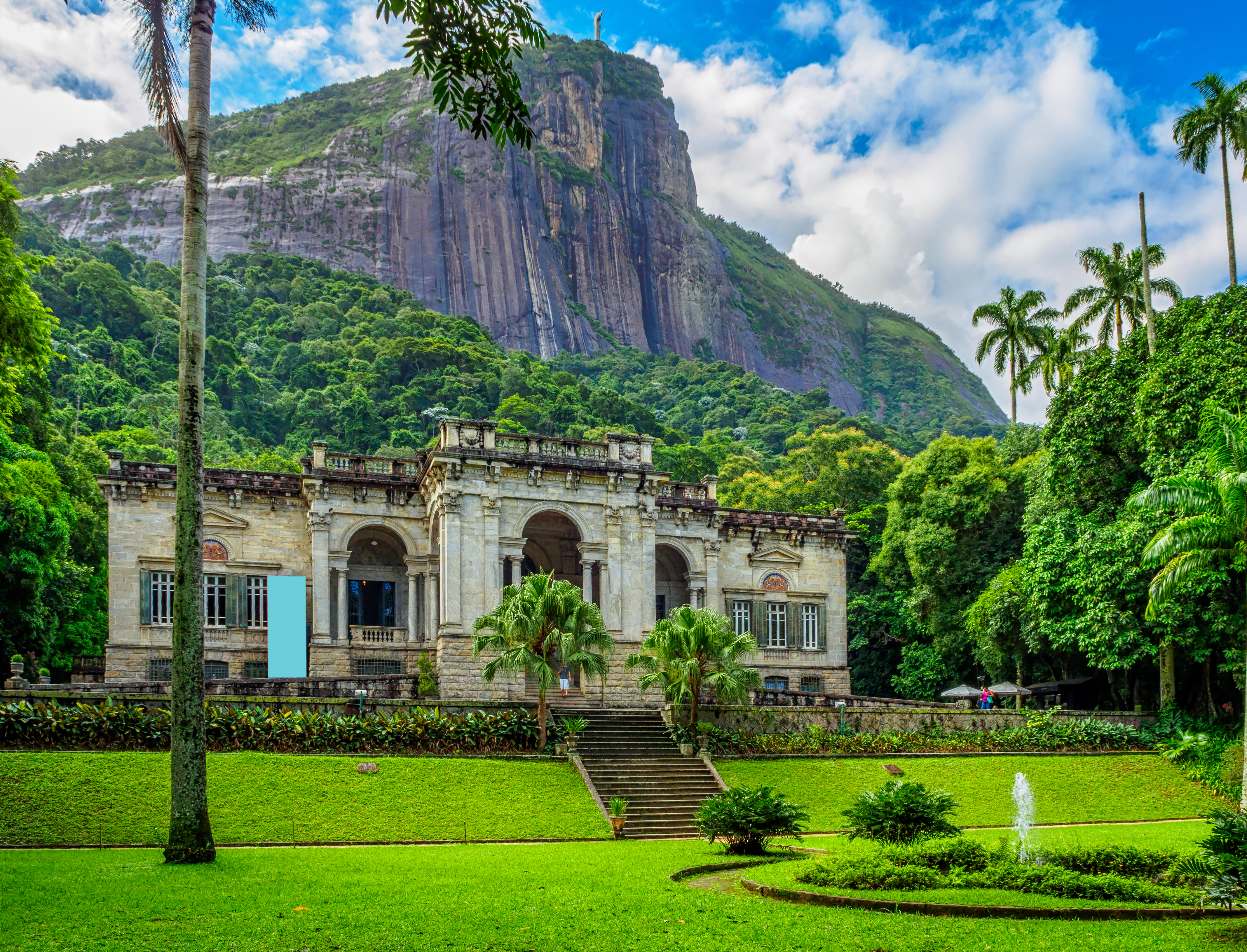 An old stone building sits at the bottom of a rocky cliff. The building, which is surrounded by leafy trees, overlooks a set of stone stairs leading down to a well-kept lawn with a fountain.