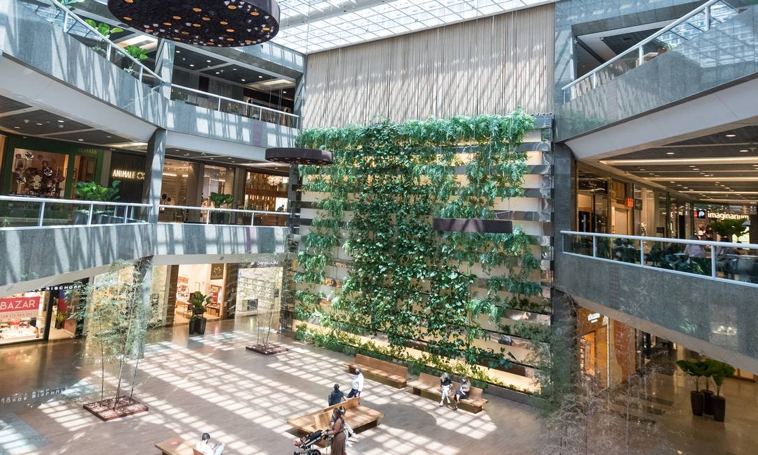 Shopping mall interior. An open space with benches and trees. Shoppers sit and chat, and wander about the space. At the rear of this area, a multistory plant wall, with ferns, ivies and other greenery. To the left and right of the plant wall and open space are three levels of shops.