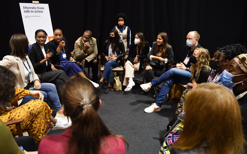 A diverse group of people sit in a circle. One person appears to be speaking, and the others appear to be listening to them. One person stands outside the circle. They appear to be listening as well. A sign on an easel to the left of the group reads 'Diversity from talk to action'.