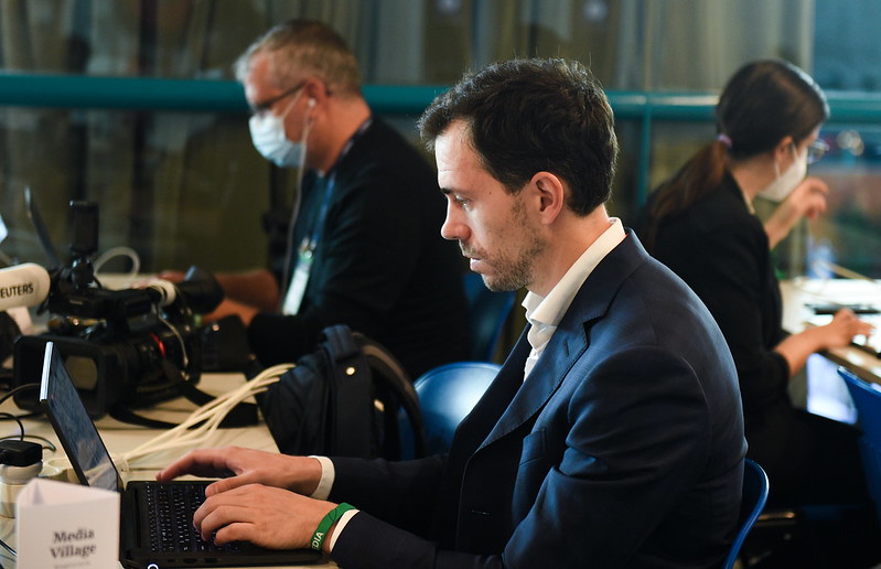 A person in a suit sits at a long desk and types on a laptop. They are viewed in profile. A small sign next to the laptop reads 'Media Village'. Two other people are visible in the background, also wearing suits and also seemingly working on laptops.