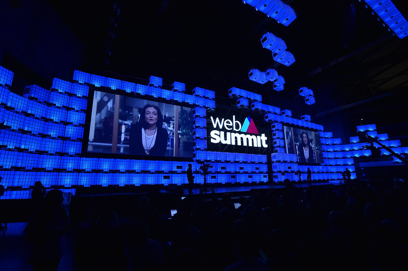 A large stage viewed over the silhouetted heads of an audience. Two large screens are on the stage, on either side of the Web Summit logo. Sheryl Sandberg, COO of Facebook, is on the screens, speaking via video link.