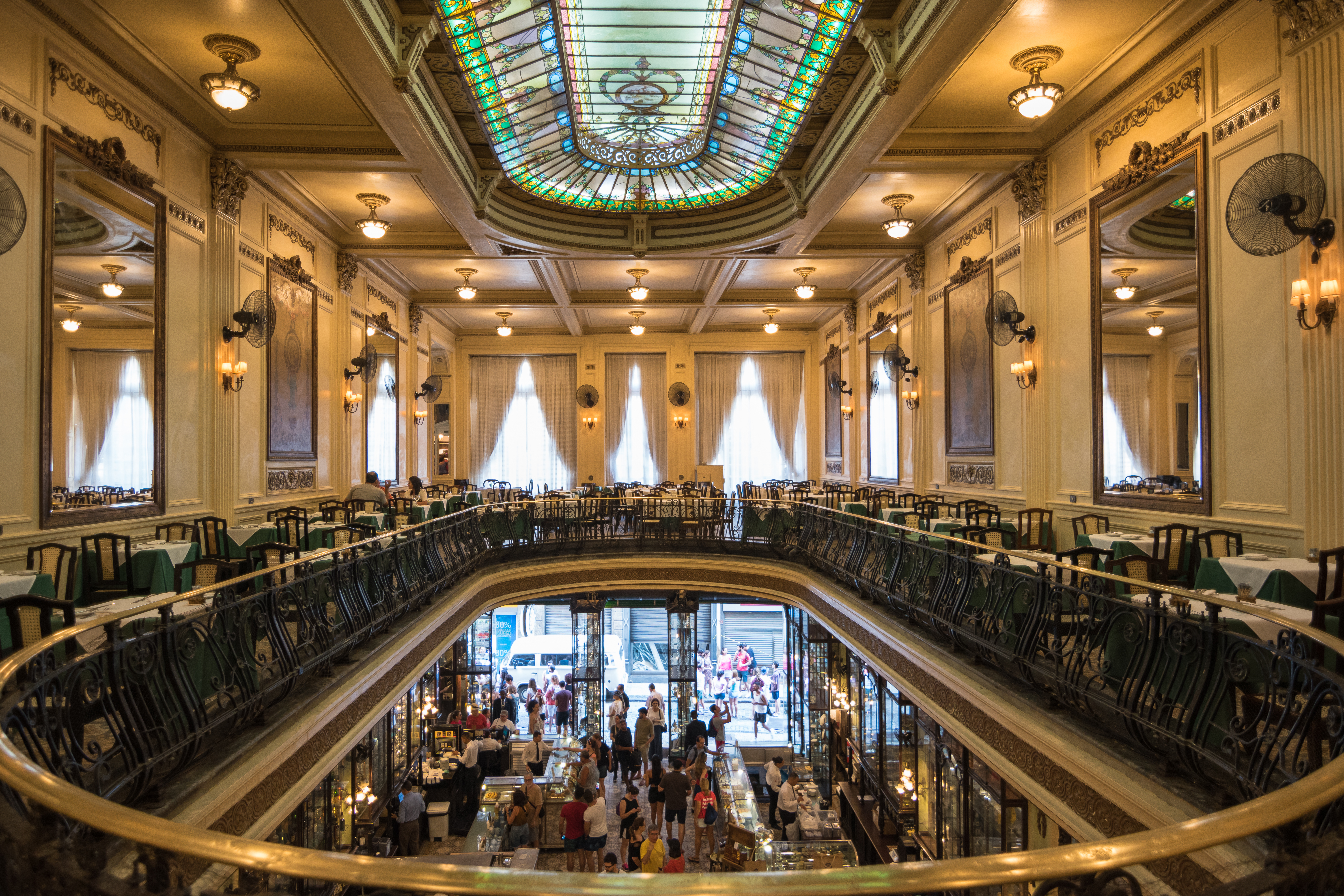 Confectionary shop and restaurant interior. An oval room with very high ceilings. A mezzanine level with a cast-iron railing wraps around the walls. A brightly coloured Victorian-style skylight is in the centre of the roof. Mirrors are hung on the walls of the mezzanine level, which is empty other than tables and chairs. The ground floor – which features several glass-fronted cabinets and glass display cases prominently – is crowded.