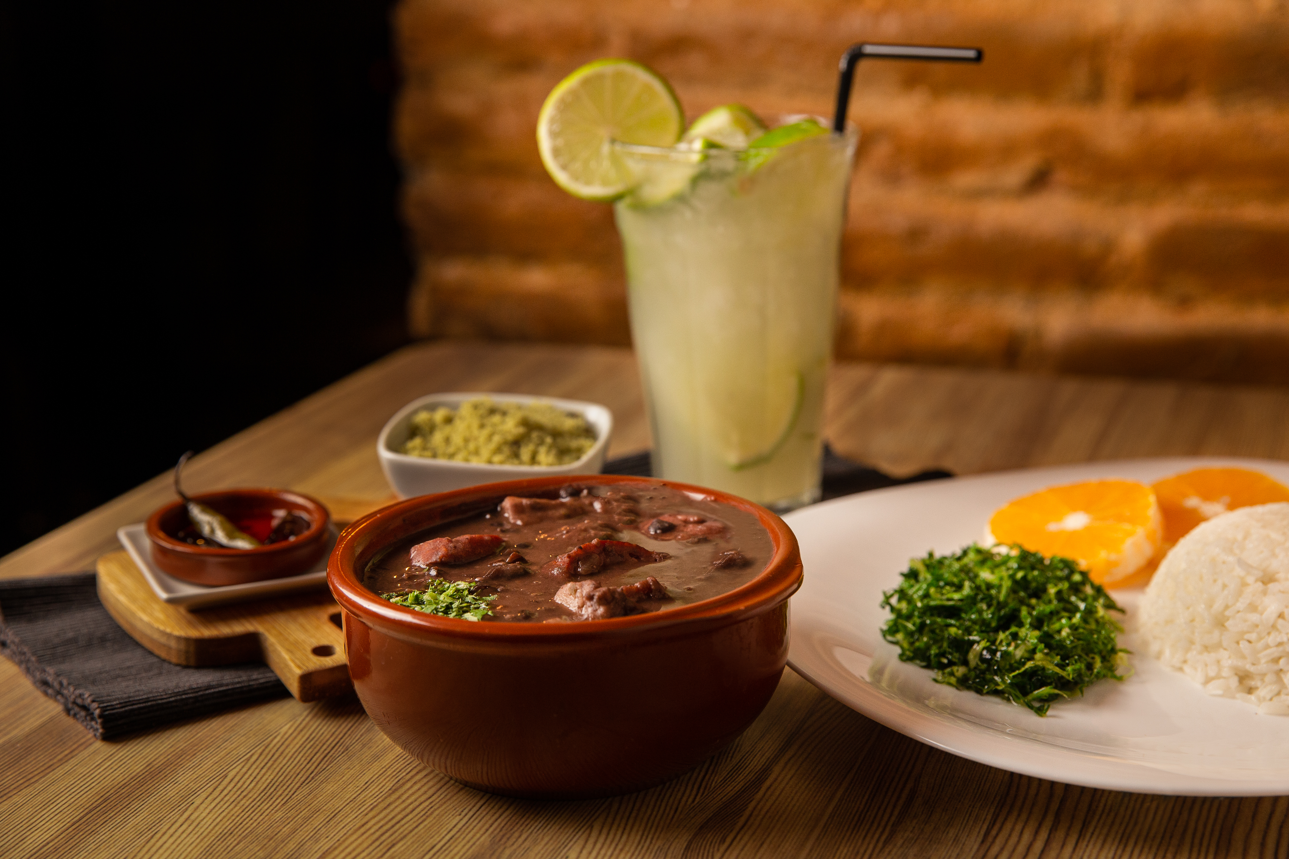 A bowl full of a traditional Brazilian dish called Feijoada – a stew containing beans, beef and pork – sits on a table. Also on the table are a plate with rice, herbs and orange slices; a dish of avocado; and a caipirinha, a drink garnished with lime.