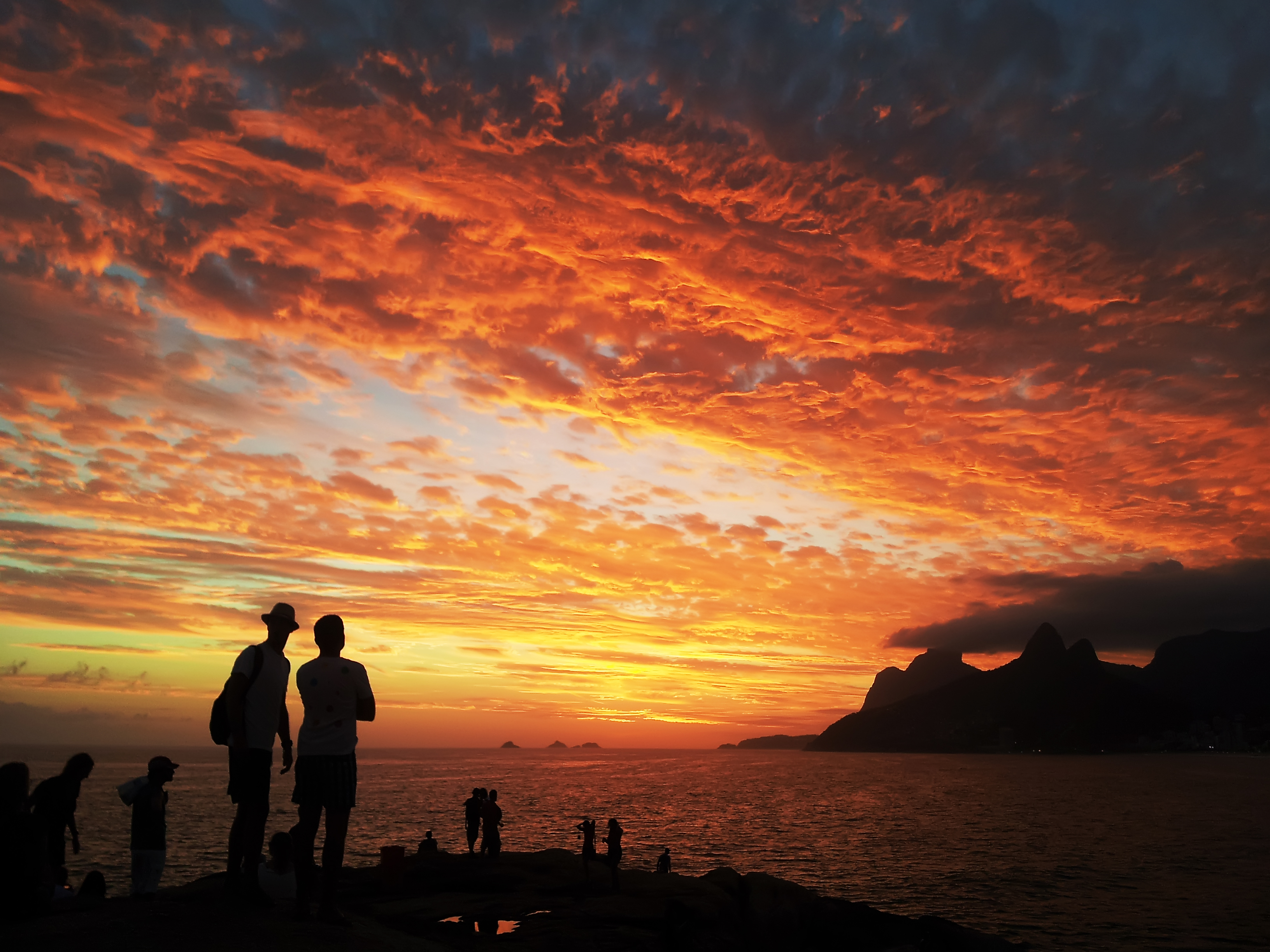 It's sunset. Several people gather on a rock next to an ocean bay. Mountains are visible on the other side of the bay.