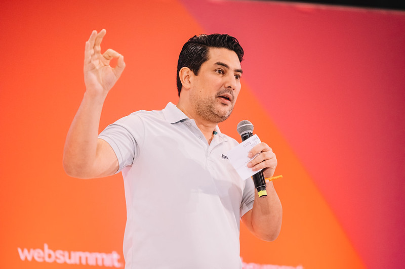 A person speaks into a hand-held microphone. A warm wall behind them features the Web Summit logo.